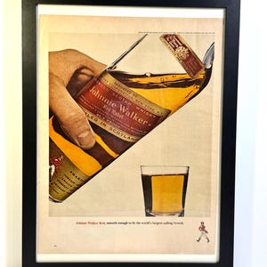 May include: Framed vintage advertisement for Johnnie Walker Red Label Scotch whisky. The image shows a bottle being poured into a glass, with the text "Johnnie Walker Red, smooth enough to be the world's largest-selling Scotch."