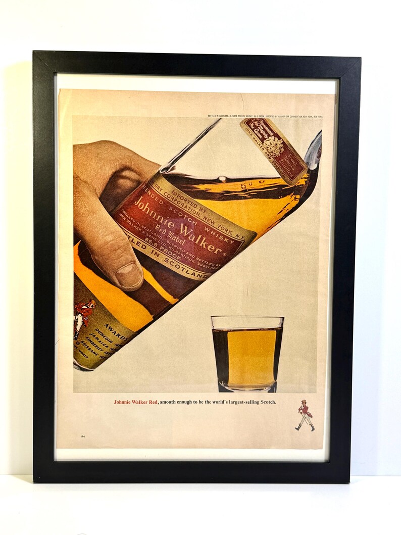 May include: Framed vintage advertisement for Johnnie Walker Red Label Scotch whisky. The image shows a bottle being poured into a glass, with the text "Johnnie Walker Red, smooth enough to be the world's largest-selling Scotch."
