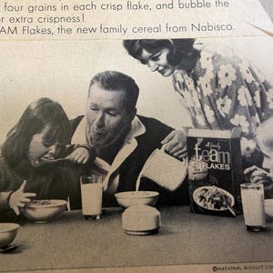 May include: Black and white vintage advertisement for Team Flakes cereal. A family is gathered around a table, eating cereal from bowls. A box of Team Flakes is visible. The text on the box reads "Team Flakes".