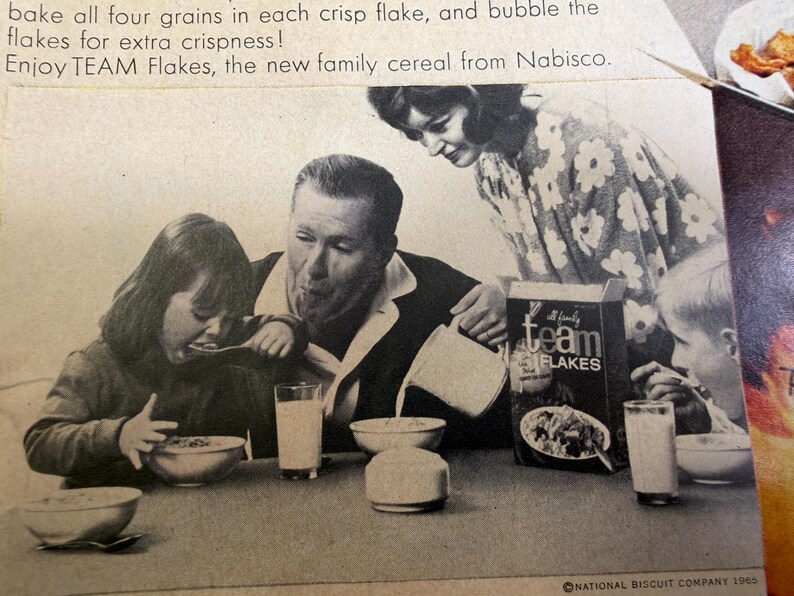 May include: Black and white vintage advertisement for Team Flakes cereal. A family is gathered around a table, eating cereal from bowls. A box of Team Flakes is visible. The text on the box reads "Team Flakes".