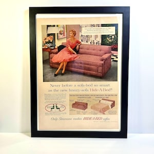 May include: Framed vintage advertisement for a Hide-A-Bed sofa. The ad features a woman in a coral dress seated on a pink sofa. Text reads, "Never before a sofa-bed so smart as the new luxury-sofa Hide-A-Bed!" and "Only Simmons makes HIDE-A-BED sofas."