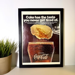 Könnte beinhalten: Gerahmte Vintage-Coca-Cola-Werbung. Die Anzeige zeigt ein Glas Cola mit Eis und eine Kugel Eis. Der Text lautet "Coke has the taste you never get tired of." Das Coca-Cola-Logo ist sichtbar. Der Rahmen ist schwarz.