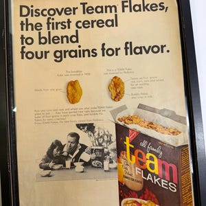 May include: Framed vintage advertisement for Team Flakes cereal. The ad features the text "Discover Team Flakes, the first cereal to blend four grains for flavor." A box of Team Flakes is shown, along with images of the cereal and a family.