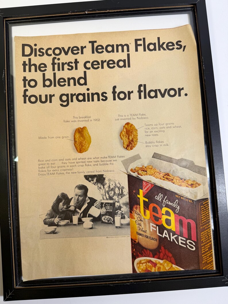 May include: Framed vintage advertisement for Team Flakes cereal. The ad features the text "Discover Team Flakes, the first cereal to blend four grains for flavor." A box of Team Flakes is shown, along with images of the cereal and a family.