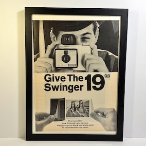 May include: Black framed vintage advertisement for the Polaroid Swinger camera. The black and white image features a man holding the camera, with text that reads "Give The Swinger 19". The ad also shows a photo of two women in graduation gowns.
