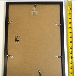 May include: A black picture frame with a brown backing. The frame is rectangular and has metal clips to hold the backing in place. A yellow measuring tape is next to the frame, showing a length of approximately 18 inches.