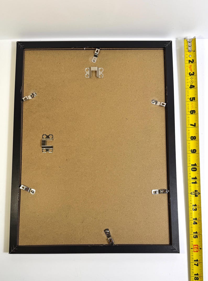 May include: A black picture frame with a brown backing. The frame is rectangular and has metal clips to hold the backing in place. A yellow measuring tape is next to the frame, showing a length of approximately 18 inches.