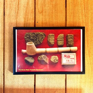 May include: A framed display featuring a wooden pipe, various green and brown substances, and a small box with Chinese text. The display is set against a red background, with the text "The Seven Steps Towards Happiness..." at the bottom.