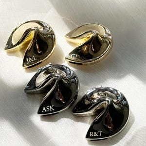 May include: Four metallic fortune cookie-shaped objects, two gold and two silver, each with engraved initials. The gold cookies have "J&L" and "GD" engraved. The silver cookies have "ASK" and "R&T" engraved. The cookies are arranged on a light-colored surface.
