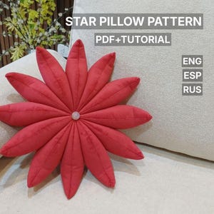 Puede incluir: Un cojín rojo en forma de estrella con un botón en el centro, colocado sobre un cojín beige. La imagen incluye el texto "STAR PILLOW PATTERN PDF+TUTORIAL" y las abreviaturas de idioma "ENG", "ESP" y "RUS".