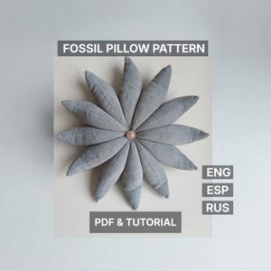Puede incluir: Un cojín gris en forma de flor con un centro rosa. El cojín está hecho de tela suave y tiene un patrón de fósil. El texto en la imagen dice "FOSSIL PILLOW PATTERN" y "PDF & TUTORIAL".