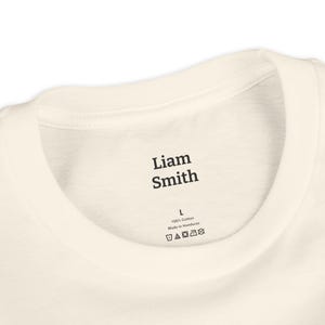 May include: Close-up of a cream-colored t-shirt collar. The text "Liam Smith" is printed in black, along with a size "L" and care instructions. The shirt is made of 100% cotton.