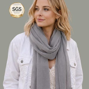 May include: A light gray scarf with fringed edges is draped around the neck. The person is wearing a white jacket and a white top. The image includes a gold and white SGS Detestet badge.