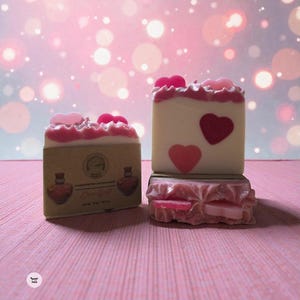 May include: Artisan soaps with heart designs. One soap is wrapped in brown paper with "Love Spell" text. The soaps are white with pink accents and heart embellishments. The background is a soft pink with bokeh lights.