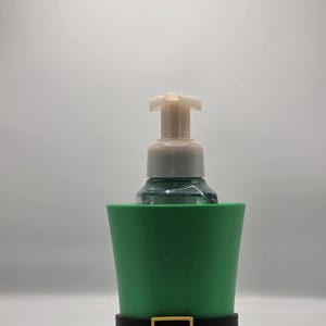 May include: A green soap dispenser in the shape of a leprechaun hat. The hat has a black band with a gold buckle. The soap dispenser is white and clear.