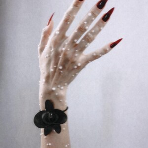 May include: A black orchid-shaped wrist accessory featuring a dark, matte flower design. The wrist and hand are adorned with numerous small, white, spherical pearl-like accents, complemented by long, pointed fingernails painted in a deep, dark red.