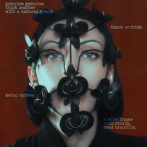 Op de afbeelding: Een zwart lederen gezichtsmasker met orchideevormige uitsparingen, metalen spikes en een draadframe met net vlechtwerk. De tekst op de afbeelding luidt: made by human hands, genuine thick leather with a natural grain, black orchids, metal spikes.