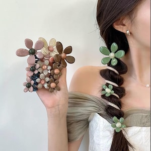 May include: A collection of flower-shaped hair accessories in various colors, including pink, brown, and green. The accessories are clustered together and also used to decorate a braid. The hair accessories are made of plastic and have a glossy finish.