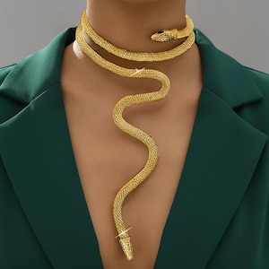 May include: A gold snake necklace with a textured, scale-like design. The necklace wraps around the neck and drapes down the chest, featuring a snake head and a pointed tail. The jewelry is worn over a dark green blazer.