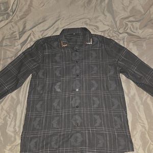 May include: A dark gray button-down shirt with a plaid pattern and long sleeves. The shirt features a collar with decorative gold-colored accents. The plaid design incorporates a repeating bat-like motif.