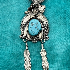 May include: A silver brooch featuring a central oval turquoise stone surrounded by embossed leaf patterns. The top displays a detailed eagle with spread wings, while the bottom features two dangling silver feathers suspended by wire wraps.