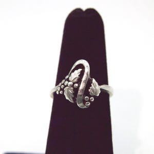 May include: A silver ring featuring a polished, curved band design accented with embossed grape leaf motifs and small clusters of round berries. The metal has a bright, reflective finish with darkened recessed areas that highlight the intricate details.