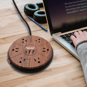 May include: A round wooden power strip with multiple outlets and USB ports. The power strip has a dark wood grain finish and a black base. The brand name "DoloBird" is printed on the surface. A laptop, headphones, and pen are in the background.