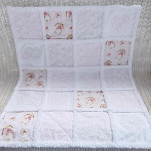 May include: White baby blanket with a patchwork design. The blanket features squares of white fabric with various textures, interspersed with squares of fabric printed with bunnies and floral patterns. Some squares have heart-shaped embroidery.