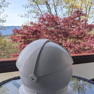 May include: A white, spherical, dome-shaped object with a smooth, matte finish. It features a central, curved panel that overlaps the main body, secured by a circular side fastener. The structure has a wide, flat base rim and a clean, minimalist design.