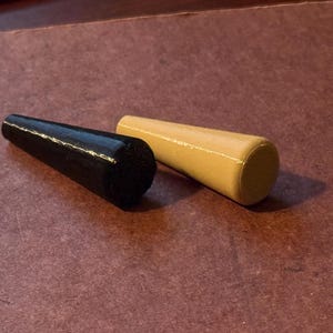 May include: Two wooden bottle stoppers. One is black, the other is light tan. Both are cylindrical with a rounded top. The stoppers are lying on a dark brown surface.