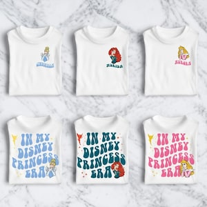 May include: White t-shirts featuring Disney princess designs. The shirts display images of Cinderella, Merida, and Aurora, along with the text "IN MY DISNEY PRINCESS ERA". The designs use blue, pink, and teal colors.