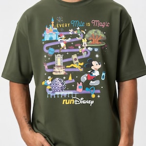 May include: Olive green t-shirt featuring a colorful graphic of Disney characters running a race. The design includes a castle, landmarks, and the text "Every Mile is Magic" and "runDisney". Mickey Mouse is prominently displayed.