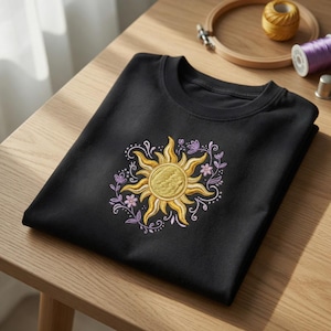 Puede incluir: Una camiseta negra con un diseño de sol bordado en amarillo y dorado, rodeado de flores y vides moradas. La camiseta está doblada sobre una superficie de madera, mostrando el intrincado bordado.