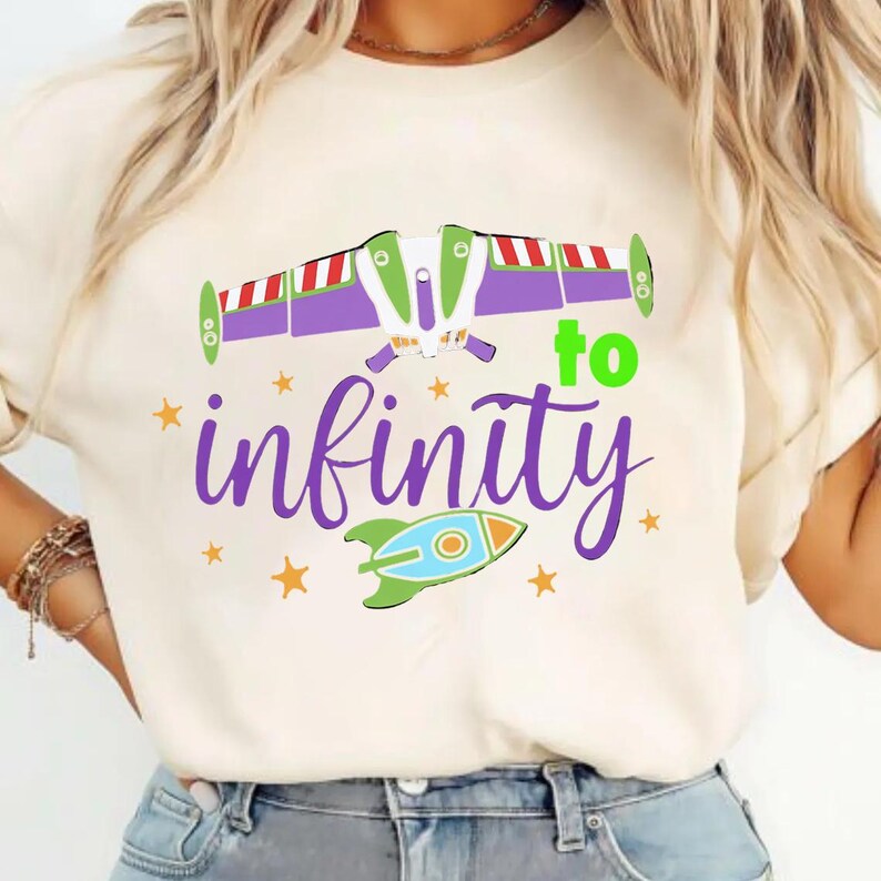 May include: Cream-colored t-shirt featuring a graphic design with the text "to infinity" in purple script. The design includes a rocket ship and a stylized spaceship with purple wings and red and white stripes. Small yellow stars are scattered around the design.