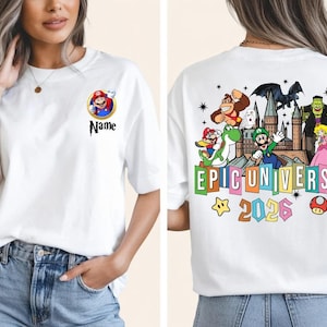 Op de afbeelding: Wit T-shirt met een kleurrijke afbeelding van verschillende stripfiguren en de tekst "EPIC UNIVERSE 2026". De voorkant van het shirt heeft een Mario-personage-ontwerp met het woord "Name" eronder. De achterkant van het shirt heeft een grote afbeelding.