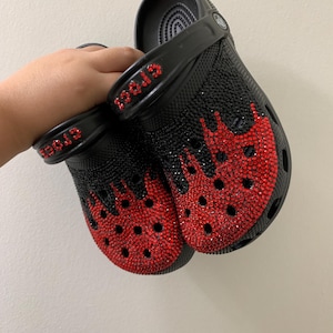 May include: Black clogs embellished with red rhinestones in a flame design. The word "CROCS" is spelled out in red rhinestones on the strap. The clogs are held up against a white background.