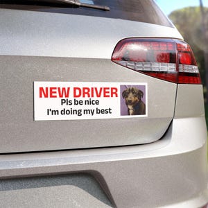Peut inclure: Un autocollant rectangulaire blanc pour pare-chocs avec le texte rouge et noir NEW DRIVER Pls be nice I'm doing my best. L'autocollant présente un encart photographique carré d'un chien noir et marron avec un collier bleu sur fond violet.