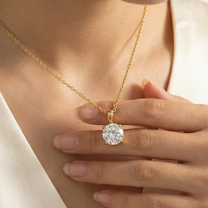 May include: A gold-colored necklace with a large, round, clear gemstone pendant. The pendant is held by a four-prong setting. The necklace is worn around the neck.