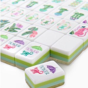 May include: A set of white Mahjong tiles with green and light blue stripes. The tiles feature various floral and animal designs, along with the word "Joker" and a monkey illustration. The tiles are arranged in a grid.