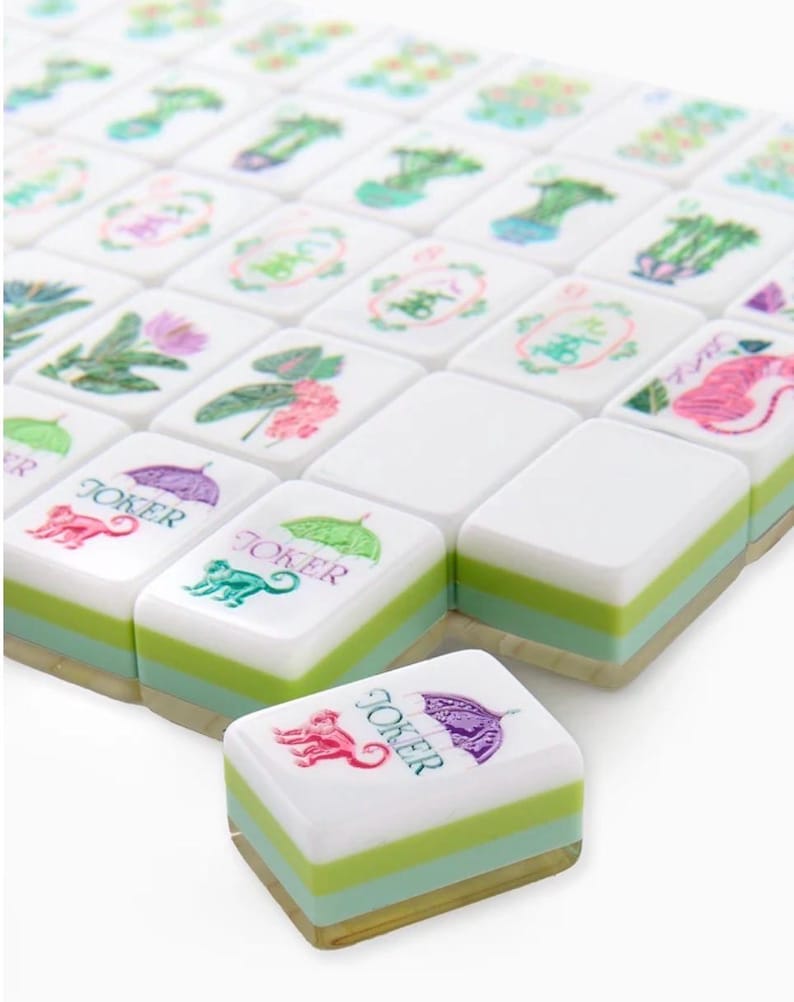 May include: A set of white Mahjong tiles with green and light blue stripes. The tiles feature various floral and animal designs, along with the word "Joker" and a monkey illustration. The tiles are arranged in a grid.