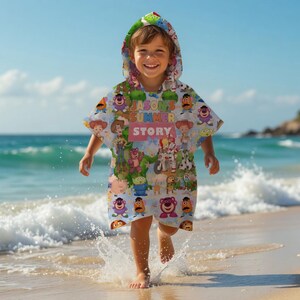 May include: A child wearing a hooded beach towel with a colorful Toy Story design. The towel features various characters from the movie and the text "JASON'S SUMMER STORY". The child is on a sandy beach near the ocean.