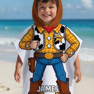 May include: A white hooded beach towel featuring a cartoon cowboy design. The design includes a yellow plaid shirt, a black and white cowhide vest, blue jeans, and brown cowboy boots. The name "JAMES" is printed on the towel.