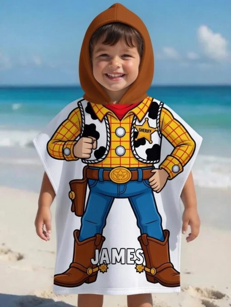 May include: A white hooded beach towel featuring a cartoon cowboy design. The design includes a yellow plaid shirt, a black and white cowhide vest, blue jeans, and brown cowboy boots. The name "JAMES" is printed on the towel.