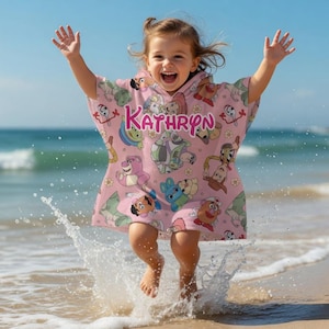 May include: A child wearing a pink hooded beach towel with cartoon character designs and the name "Kathryn" in pink letters. The child is jumping in the air on a sandy beach, with water splashing around their feet.