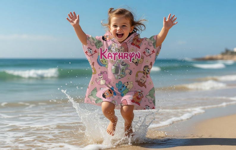 May include: A child wearing a pink hooded beach towel with cartoon character designs and the name "Kathryn" in pink letters. The child is jumping in the air on a sandy beach, with water splashing around their feet.