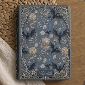 Pode incluir: Um diário de capa dura azul com uma borda dourada intrincada, um padrão repetitivo de dragões pretos e ilustrações florais em tom creme. O nome Meredith está gravado numa moldura decorativa na parte inferior central da capa.