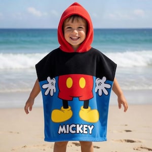 K&ouml;nnte beinhalten: Ein Kind tr&auml;gt ein Kapuzen-Strandtuch mit einem Mickey Mouse-Design. Das Handtuch hat eine rote Kapuze, einen schwarzen oberen Bereich und einen blauen unteren Bereich mit Mickeys Bild und dem Wort "Mickey". Das Kind befindet sich an einem Sandstrand mit dem Meer im Hintergrund.