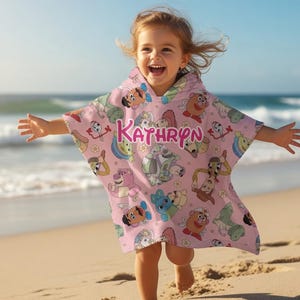 May include: A pink hooded beach towel with cartoon character graphics and the name "Kathryn" in pink letters. The towel is being worn by a child on a sandy beach with the ocean in the background.