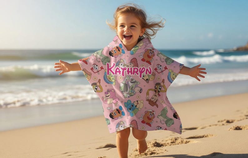 May include: A pink hooded beach towel with cartoon character graphics and the name "Kathryn" in pink letters. The towel is being worn by a child on a sandy beach with the ocean in the background.
