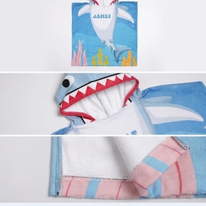 K&ouml;nnte beinhalten: Blaues Handtuch mit Kapuze im Hai-Design mit dem Namen "JAMES" auf der Vorderseite. Das Handtuch hat eine Haifischkopf-Kapuze mit roten und wei&szlig;en Z&auml;hnen. Das Design umfasst eine Hai-Illustration und Unterwasserelemente.